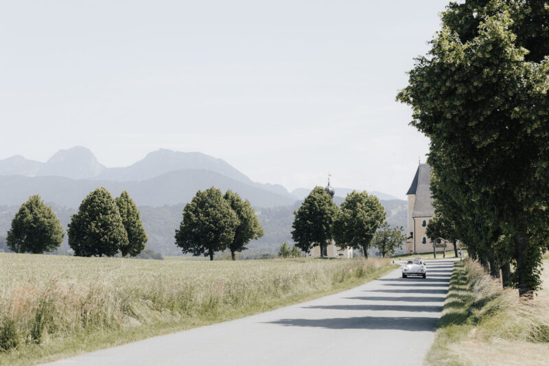 Brautpaar in einem alten Käfer mit Alpen und Kirche Wilparting im Hintergrund