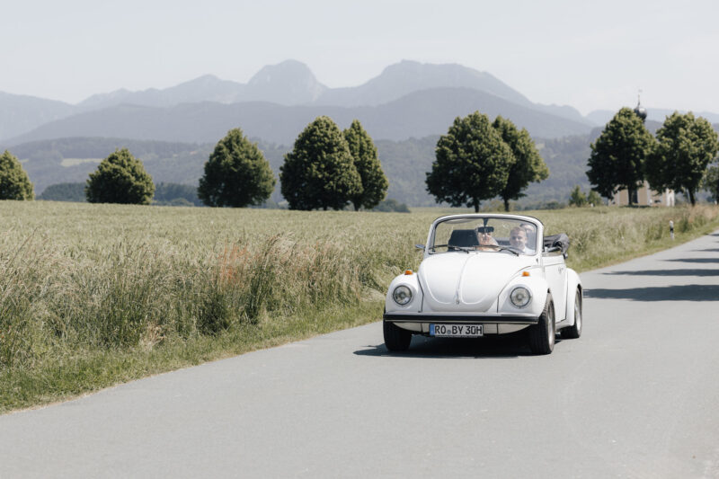 Brautpaar in einem alten Käfer mit Alpen und Kirche Wilparting im Hintergrund